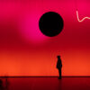 Vor einer großen, in Rottönen leuchtenden Projektionsfläche zeichnet sich die dunkle Silhouette eines Performers ab, über ihm schwebt eine schwarze Scheibe.