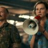 A woman with a megaphone looks out of the picture frame in horror. She is wearing a loose denim shirt and has shoulder-length hair. Next to her stands a man in military uniform and a moustache.