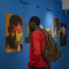 A Black man with a red shirt and a grey backpack is looking at a photo exhibition on a bright blue wall. In one photo, a Black person has a rainbow-coloured heart painted on his face.