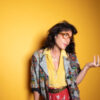 Joan As Police Woman stands in front of a yellow background and wears a combo of yellow blouse, red pants and patterned jacket, plus large glasses, her left hand denotes a half questioning, half challenging gesture.
