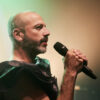 Jan Plewka, a white man with a bald head and short beard, holds a microphone with both hands and sings tenderly into it. He wears red lipstick and a black top. The background is lightly illuminated in green.