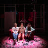 Three young people sit on a red sofa with a package. They look gloomy while confetti is snowing down on them. Behind them is a spatial construction in which two other people sit in the red light.