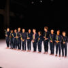 12 Tänzerinnen in dunkelblauen Anzügen stehe auf der Bühne auf weißem Tanzboden. Sie habe alle eine Blume in der Hand.