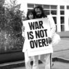 Schwarz-weiß Foto von einer Frau und einem Mann, die weiß gekleidet und mit langen offenen Haaren auf einem Paar Stufen draußen stehen und ein großes Schild halten: WAR IS NOT OVER. Im Hintergrund eine große Topfpflanze.