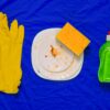 On a bright blue cloth lie a pair of yellow rubber gloves, a dirty white porcelain plate with a yellow sponge and a green washing-up liquid bottle.