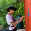 A woman in a white T-shirt and blue hat paints a blue bird on an orange wall with a brush and looks concentrated while working. Dense green bushes grow behind her.
