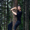A slender white woman in tight black pants and blouse clutches a tree trunk in the forest. She hangs from the tree, her arms and legs wrapped around the trunk. She is barefoot and has her dark hair tied back. The forest is dense and green.