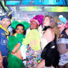 Five performers wear garishly colored, wildly patterned costumes and makeup and stand with expressive facial expressions sideways in front of a wall painted with colorful graffiti.