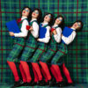 Five women in green Scottish tartan dresses, white blouses, red tights and bright blue eye shadow stand closely in a row in front of a background in the pattern like their dresses. They hold blue clipboards and red cups in their hands.