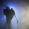 A bald man wears a feather boa and stands sideways to the camera in the fog at a microphone stand. He sings and is illuminated by blue light from behind.