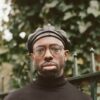 Ghostpoet ist a Black Man with round glasses and a short beard. He wears a black beret and a tight black turtleneck. In the background is an elegant green metal fence and a tree.