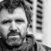 Schwarz-weißes-Portraitfoto einer männlich gelesenen Person mittleren Alters mit Bart. Sie trägt einen Schal, mehrere Ohrringe im einen Ohr und schaut ernst.