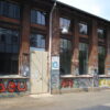 The Kampnagel main building from behind: An old brick factory building sprayed with graffiti. On a large gray steel door a sign with "K3" in turquoise letters.