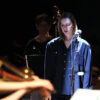 A young white woman stands in front of a microphone with her eyes downcast. She is wearing an oversized blue shirt with gold buttons. She is half-lit by a beam of light. In front of her sits a blurred violinist. Behind her, a cellist in the dark.