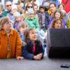 Strahlende und staunende Kinder in bunten Jacken stehen vor einer Bühne.