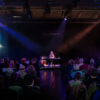 Ein Foto von einem Konzert: Eine Musikerin sitzt zentral auf der Bühne an einem Keyboad und wird von drei bunten Lichtspots angestrahlt. Das Publikum sitzt mit dem Rücken zur Kamera.
