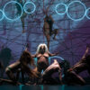 15 performers move partially or almost naked on and around a rope construction. Some wear long-haired wigs and animal-like masks. The scene is lit in dark purple and blue neon light circles hang from above.