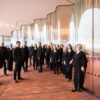 The NDR Vokalensemble, a large group of singers dressed in black, stands on the plaza of Hamburg's Elbphilharmonie in front of a corrugated glass wall.