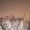 Twelve dancers in white, semi-transparent dresses with puffed sleeves, ruffles and pleats dance loosely and ecstatically with their eyes closed. Hair swings around, arms up and to the side.
