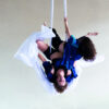 Two white people hang in the air overhead in white acrobatic scarves. They are wearing black underwear and airy blue shirts. Part of the cloth swings in the air.