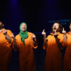 Vier Personen in orangenen Overalls stehen lachend in einem dunklen Raum und strecken jeweils beide Mittelfinger aus.