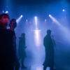 A handful of people in coats and masks roam through stage fog in a blue-lit hall. A few white spotlights shine on the floor between the people.