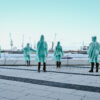 Seven people in turquoise plastic rain capes stand outside with their backs to the camera and look out over Hamburg harbour and the cranes. The sky is soft blue-white.