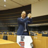 A slim white man with short blond hair in a blue suit, stands in a hall of the European Parliament with his hands folded next to his head in a winning pose. On his chair hangs a bath towel with the logo of DIE PARTEI.