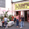 Some people are standing in small groups on the Kampnagel Piazza in front of the main entrance. There is a big yellow sign with the Kampnagel lettering in black letters. On the piazza there are light grey concrete blocks to sit on and a few palm trees.