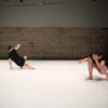 Zwei Tänzer*innen in enger schwarzer Kleidung mit kurzen Ärmeln und Hosen bewegen sich hockend auf einem weißen Tanzboden. Im Hintergrund eine unverputzte Steinwand.