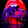 Sechs Personen in Sportkleidung im 80er-Jahre Stil posieren um einen Tresen in lila und rotem Neonlicht herum. Sie tragen fluoreszierendes Make-Up, im Hintergrund ist ein ca. 3 Meter großer knallroter Mund mit offenen Lippen und zwei Schneidezähnen.