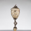 An antique-looking object made of yellowed glass in the form of bubbles and metal plant elements, reminiscent of a goblet or a lamp, with centipedes and worms crawling on it.