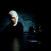 In einem dunklen Bühnenraums steht eine Person vor einem Tisch mit vielen elektronischen Musikgeräten und Computern. Daneben wird das Bild einer Figur projiziert, die sehr ausgeprägte Wangenknochen, eine Glatze und Glitzersteine statt Augenbrauen hat.