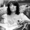 Black and white photo of the protagonist, she has a dark fringe hairstyle, wears a striped blouse and sits watching TV and folding laundry in a patterned wing chair.