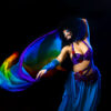 A belly dancer in a bright blue costume, a black Afro haircut and a full black beard swings a rainbow-coloured scarf behind her, smiling and elegant.