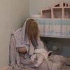 Eine Person mit langen Haaren sitzt mit einem Baby auf dem Boden neben einem Tisch mit einem Modellhaus.