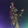 A drag queen with green make-up on her face, a red hat and a green-brown costume stands with her hands raised between purple and green beams of light.