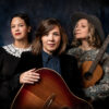 Vor einem dunklen Hintergrund stehen drei Frauen mit eleganter Kleidung. Zwei halten eine Gitarre vor sich.