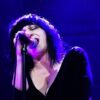 Lydia Lunch wears a wide-cut black dress and sings into a microphone with her eyes closed; the image is bathed in purple-blue light.