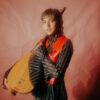 A young woman with short brown hair holds a light brown Bağlama next to her. She wears a red waistcoat over a dark striped blouse with wide sleeves and has red eye shadow....