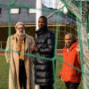 An older white man and two younger black men in winter jackets and scarves stand in a football goal on bright green turf. They look seriously into the camera, one holding onto the net.