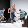 On a white stage floor, three people in rubber boots and everyday clothes shovel earth into piles.