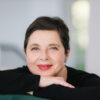 Isabella Rosellini, a white older woman with short dark hair and red lipstick, wears a black long-sleeved top and rests her chin on her bent arms on the back of a chair in front of her. She smiles gently.