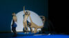Vier Performer*innen in eng anliegenden, beigen Kostümen stretchen sich auf einem blauen Tanzboden vor einem schwarzen Hintergrund mit einem hellen Lichtspot.