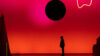 Vor einer großen, in Rottönen leuchtenden Projektionsfläche zeichnet sich die dunkle Silhouette eines Performers ab, über ihm schwebt eine schwarze Scheibe.