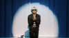 Ein junger Mann mit weißer Perücke und schwarzer Sonnenbrille à la Andy Warhol kniet in betender Haltung vor einem blauen Vorhang, ein heller Lichtspot ist auf ihn gerichtet.