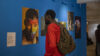 Ein Schwarzer Mann mit rotem Hemd und grauem Rucksack betrachtet eine Fotoausstellung an einer knallblauen Wand. Auf einem Foto ist eine Schwarze Person mit einem regenbogenfarbenen Herz aufs Gesicht gemalt.