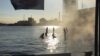 In einem Hafenbecken zeichnen sich die Silhouetten mehrerer Frauen gegen das Sonnenlicht ab, es scheint so, als würden sie auf dem Wasser laufen.
