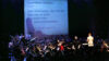 Ein Orchester spielt auf einer Bühne, im Hintergrund ist eine große Leinwand, auf die ein Songtext projiziert wird. Vor dem Orchester steht eine junge Frau, die fröhlich in ein Mikrofon singt.
