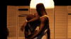 Zwei Schwarze Männer mit kurzen Haaren in Unterhosen und weißen Unterhemden stehen in einer kunstvollen Bewegung zärtlich ineinander verschlungen vor einem Bett mit rotem Bezug und mit einem hölzernen Raumtrenner dahinter. Die Szene ist dunkel beleuchtet.